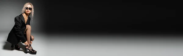Donna seducente ed elegante in occhiali da sole scuri, blazer e calze che fissano sandalo tacco e distogliendo lo sguardo su sfondo nero e grigio, moda sexy, banner con spazio copia — Foto stock