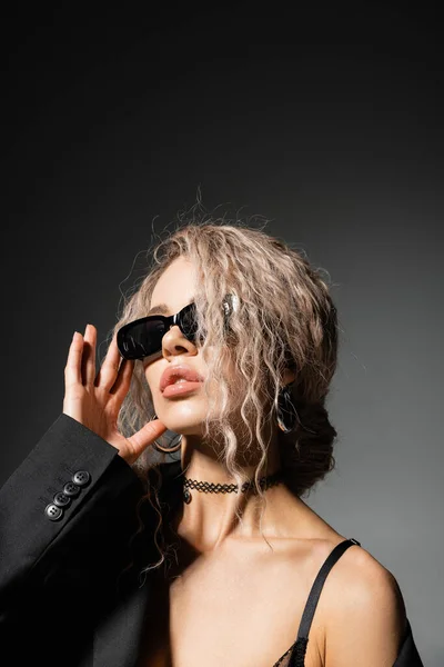 Retrato de mujer sexy en chaqueta y collar ajustando gafas de sol oscuras y mirando hacia otro lado sobre fondo negro y gris, pelo rubio ceniza, estilo glamour, confianza, auto-expresión moderna - foto de stock