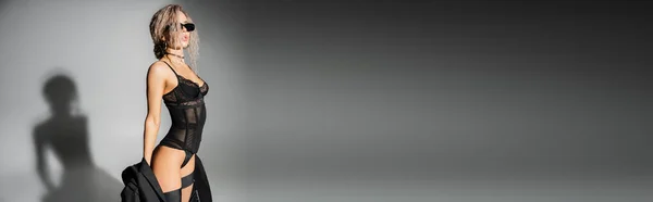 Mujer elegante y sexy con pelo rubio de ceniza ondulada, en gafas de sol oscuras, body negro y medias que sostienen la chaqueta mientras están de pie sobre fondo gris, moda y sexualidad, pancarta con espacio para copiar - foto de stock