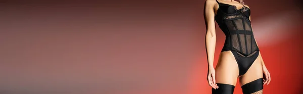 Обрезанный вид сексуальной женщины модели с тонким телом в черном телесном костюме и чулках, стоя на красном и сером фоне, чувственность и мода, баннер с копировальным пространством — стоковое фото