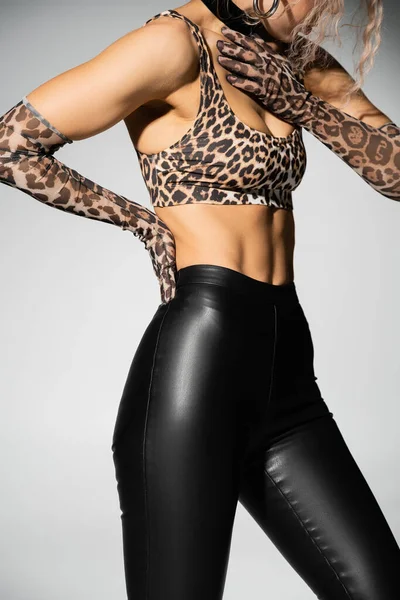 Sexuality and style, cropped view of slender woman in black latex pants, animal print crop top and long gloves posing with hand on waist on grey background, modern fashion trend — Stock Photo