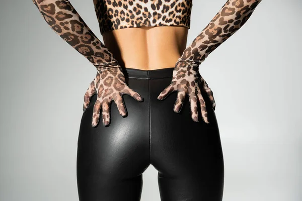 Обрезанный вид на модную женщину в перчатках для печати животных, топ и черные латексные брюки, трогающие сексуальные ягодицы и позирующие на сером фоне, современная индивидуальность, самовыражение — стоковое фото