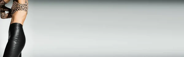 Individualidade moderna, olhar sexy, vista cortada de modelo feminino esbelto em pé em calças de látex preto, top de colheita de impressão animal e luvas longas em fundo cinza, banner com espaço de cópia — Fotografia de Stock