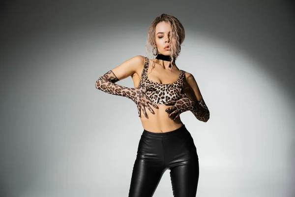 Autoexpresión moderna, mujer apasionada y glamurosa en top estampado de leopardo, guantes largos y pantalones de látex negro tocando cuerpo delgado sobre fondo gris, fotografía sexy de moda - foto de stock