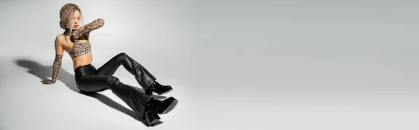 Modische und sexy Frau in schwarzer Latexhose, bauchfreiem Leopardenmuster und langen Handschuhen auf grauem Hintergrund, schlanker Körper, welliges aschblondes Haar, Banner mit Kopierplatz — Stockfoto