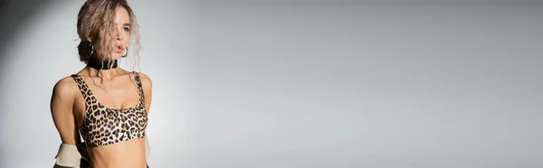 Modello femminile moderno e sensuale in top crop top stampa leopardo in piedi e distogliendo lo sguardo su sfondo grigio, corpo snello, capelli biondi cenere ondulati, fotografia di moda sexy, banner con spazio copia — Foto stock