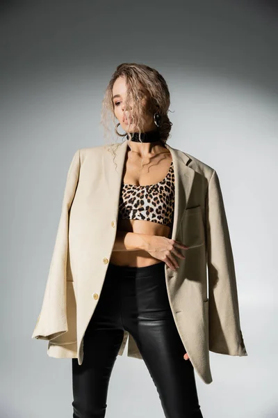 Sensuality and fashion, charming woman with dyed ash blonde hair and slender body posing in animal print crop top and black latex pants, with beige blazer on shoulders on grey background — Stock Photo