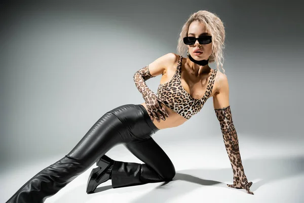 Mujer de moda con cuerpo sexy y pelo rubio ceniza teñido posando en gafas de sol oscuras, top de estampado animal, guantes largos y pantalones de látex negro sobre fondo gris, auto-expresión moderna - foto de stock