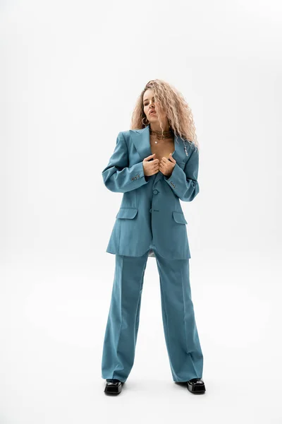 Full length of appealing and seductive woman with wavy ash blonde hair standing in blue oversize blazer and pants while looking away on grey background, modern personality, stylish look — Stock Photo