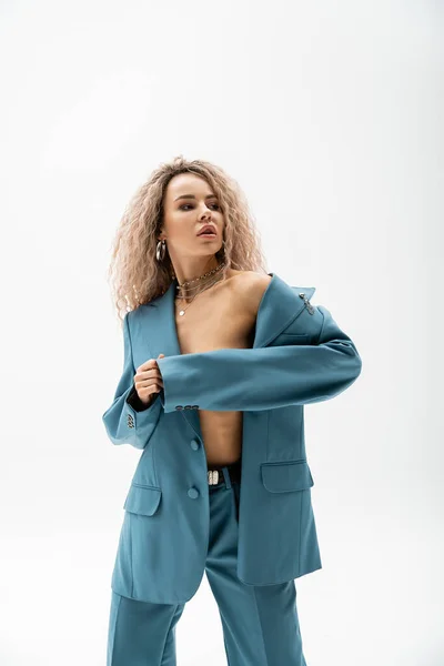 Sexuality and fashion, seductive woman with wavy ash blonde hair looking away while wearing blue oversize suit on shirtless body and posing on grey background, expressive individuality — Stock Photo