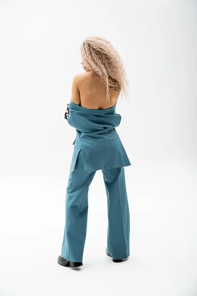 Back view of seductive and tattooed woman with bare shoulders wearing blue oversize pantsuit on shirtless body while standing on grey background, glamour fashion, modern self-expression — Stock Photo