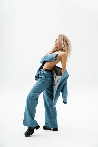 Longitud completa de la mujer expresiva y de moda que cubre el pecho desnudo con el brazo mientras posa en traje azul de gran tamaño sobre el cuerpo sin camisa sobre fondo gris, estilo glamour, vista lateral - foto de stock