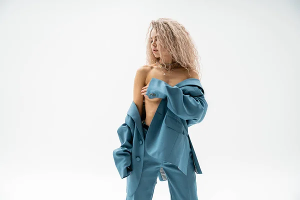 Sexy fashion trend, erotic woman with dyed and wavy ash blonde hair covering naked breast with hand and standing in blue oversize pantsuit on grey background, glamour and self-expression — Stock Photo