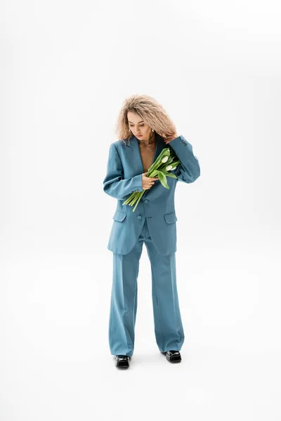 Femininity and style, full length of stylish and beautiful woman with dyed ash blonde hair holding bouquet of white tulips while standing in blue oversize suit on grey background — Stock Photo