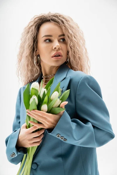 Retrato de mujer de moda con mirada expresiva posando con ramo de tulipanes blancos y mirando hacia otro lado sobre fondo gris, pelo rubio de ceniza ondulada, chaqueta azul de gran tamaño, sesión de fotos de moda - foto de stock