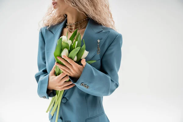 Vue partielle de la femme à la mode en blazer oversize bleu tenant bouquet de tulipes blanches debout sur fond gris, cheveux blonds cendrés ondulés, accessoires argentés, style moderne — Photo de stock