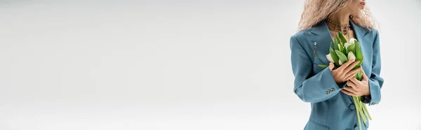 Vista ritagliata di donna alla moda con capelli biondi cenere ondulati con bouquet di tulipani bianchi mentre in piedi in tuta oversize blu su sfondo grigio, banner con spazio copia, sensualità e moda — Foto stock