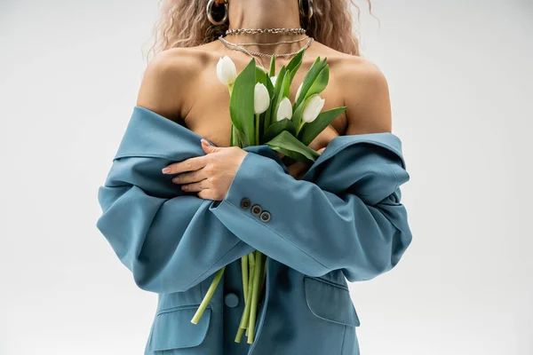 Partial view of sexy woman with silver necklaces and wavy ash blonde hair wearing blue oversize blazer on bare shoulders and holding bouquet of white tulips on grey background — Stock Photo