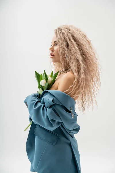 Side view of sexy woman with bare shoulders and wavy ash blonde hair wearing blue oversize blazer, holding bouquet of white tulips while standing and looking away on grey background, glamour style — Stock Photo