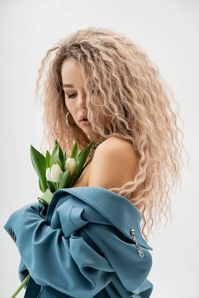 Sensual and charming woman with bare shoulders and wavy ash blonde hair holding bouquet of white tulips and posing in blue oversize blazer on grey background, modern fashion and style — Stock Photo