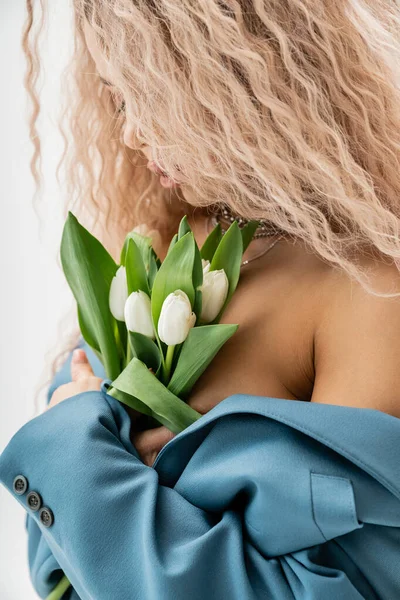 Sexualidad y moda, mujer encantadora y romántica con la ceniza ondulada pelo rubio con chaqueta azul de gran tamaño en el cuerpo sin camisa y abrazando ramo de tulipanes blancos sobre fondo gris - foto de stock