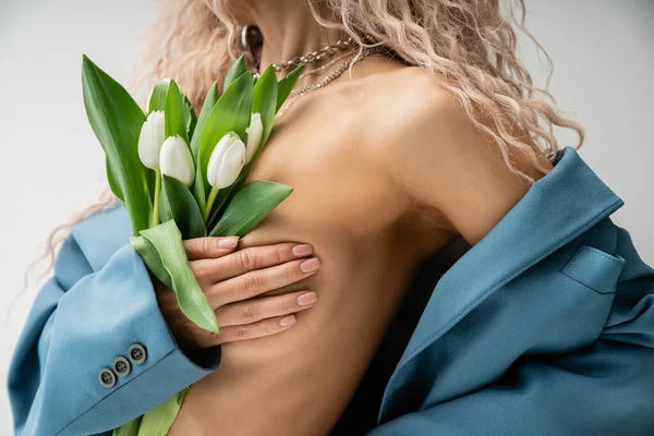 Vista cortada de mulher sexy em colares de prata cobrindo peito nu com as mãos enquanto posando com blazer azul oversize e buquê de tulipas em fundo cinza, feminilidade e moda — Fotografia de Stock