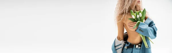 Vista cortada de mulher apaixonada e sexy com corpo sem camisa decolando blazer azul, obscurecendo o peito nu com a mão e segurando tulipas brancas no fundo cinza, banner com espaço de cópia — Fotografia de Stock