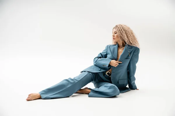 Pleine longueur de femme séduisante et pieds nus avec des cheveux blonds frêne ondulé assis en costume oversize bleu sur fond gris, représentation de la mode moderne, l'individualité et le style — Photo de stock