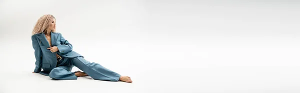 Piena lunghezza del modello attraente e alla moda a piedi nudi seduto in tuta oversize blu su sfondo grigio, capelli biondi cenere ondulato, glamour e sensualità, tendenza moda moderna, banner con spazio copia — Foto stock