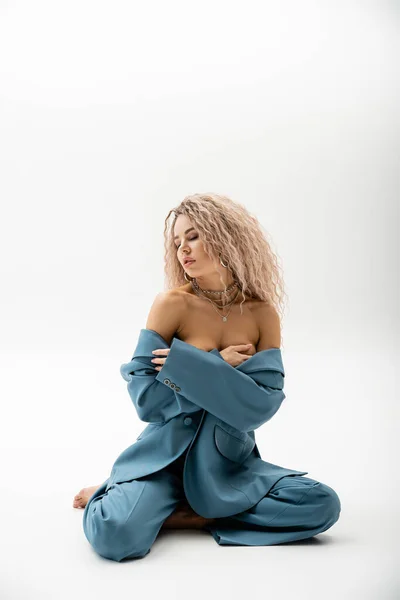 Full length of passionate woman with bare shoulders, wavy ash blonde hair and closed eyes sitting in blue oversize suit on grey background, sexy look, fashion and sensuality — Stock Photo