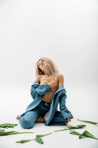 Sexy fashion photography, full length of passionate woman with dyed ash blonde hair and closed eyes covering naked breast with hand while sitting in blue oversize suit near tulips on grey background — Stock Photo