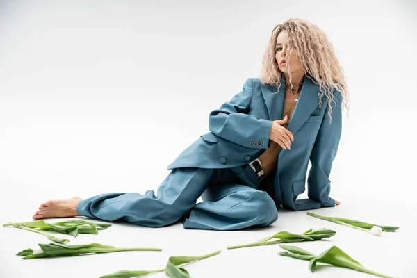 Full length of stylish and sexy female model in blue oversize suit, with expressive gaze and wavy ash blonde hair looking at camera while sitting near tulips on grey background — Stock Photo