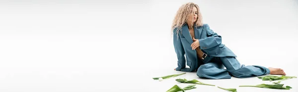 Full length of barefoot and stylish female model with wavy ash blonde hair posing in blue oversize suit while sitting near tulips on grey background, banner with copy space — Stock Photo