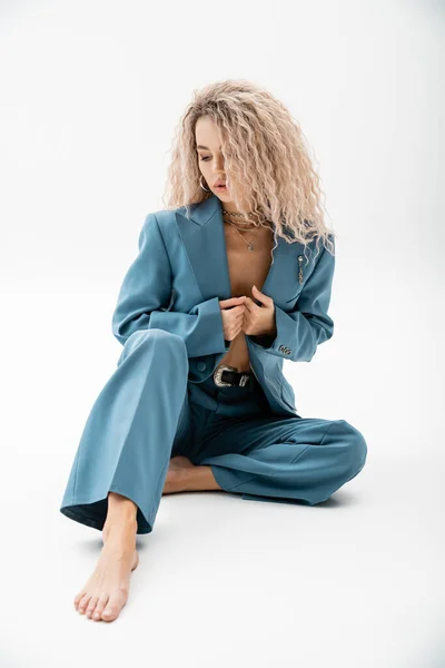 Full length of glamour and barefoot woman with dyed ash blonde hair, in blue oversize suit and silver accessories sitting on grey background, fashion-forward, modern individuality, self-expression — Stock Photo