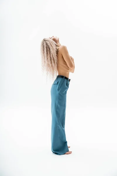 Сексуальность и стиль, полная длина соблазнительной и татуированной женщины с волнистыми светлыми волосами ясеня и безрубашечным телом, позирующим босиком в синих брюках большого размера на сером фоне, вид сбоку — стоковое фото