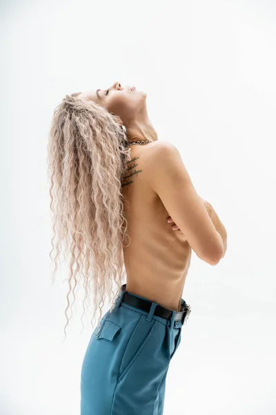 Side view of sensual woman with shirtless tattooed body and wavy ash blonde hair wearing blue oversize pants while standing with closed eyes on grey background, sexy look, self-expression — Stock Photo