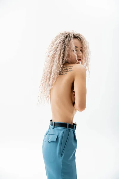 Seductive and expressive woman with shirtless tattooed body obscuring naked breast with hands while standing in blue oversize pants on grey background, fashion and sexuality, modern individuality — Stock Photo