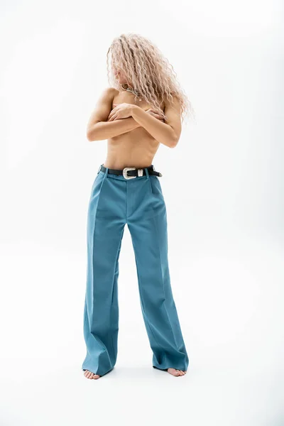 Full length of sexy barefoot woman with shirtless body and face obscured with wavy ash blonde hair posing in blue oversize pants while covering naked breast with hands on grey background — Stock Photo