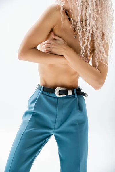 Partial view of seductive woman with shirtless body and wavy ash blonde hair obscuring naked breast with hands while posing in silver necklaces and blue pants with leather belt on grey background — Stock Photo