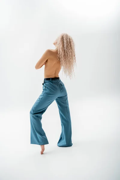 Pleine longueur de femme sexy, torse nu et pieds nus avec des cheveux blonds cendres teints posant en pantalon oversize bleu sur fond gris, corps mince, individualité et expression de soi — Photo de stock