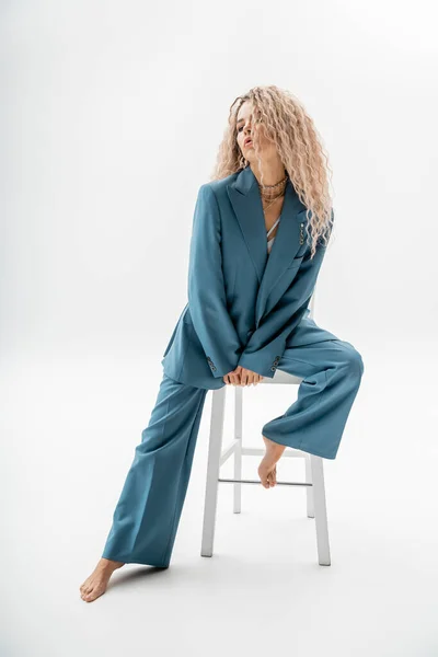 Full length of seductive, fashionable and barefoot woman with dyed ash blonde hair sitting on chair in expressive pose on grey background, sensuality, femininity and style — Stock Photo