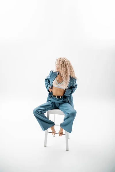 Full length of stunning barefoot woman with wavy ash blonde hair, in blue oversize suit and bra sitting on chair while posing on grey background, sexy look, modern female model, self-expression — Stock Photo