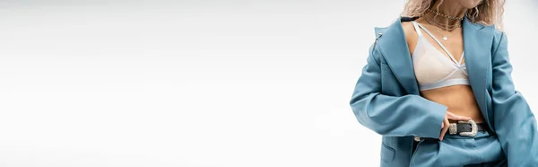 Vue partielle de femme séduisante en colliers argentés, soutien-gorge et combinaison oversize bleue touchant la ceinture en cuir tout en se tenant debout sur fond gris, look sexy, mode moderne, bannière avec espace de copie — Photo de stock
