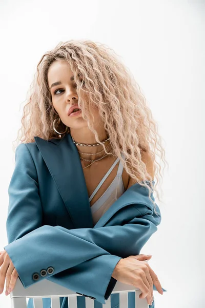 Portrait of charming woman with dyed ash blonde hair and silver accessories looking away while posing on chair in bra and blue oversize blazer on grey background, sensuality and fashion — Stock Photo