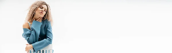 Sexy e sensuale donna in blu oversize blazer toccante collo mentre seduta su sedia con gli occhi chiusi su sfondo grigio, ondulato cenere capelli biondi, femminilità e moda, banner con spazio copia — Foto stock