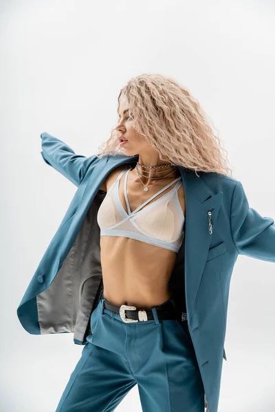 Expressive and stylish woman with trendy silver accessories and wavy ash blonde hair wearing bra, blue oversize suit and posing with outstretched hands on grey background, sexuality and fashion — Stock Photo