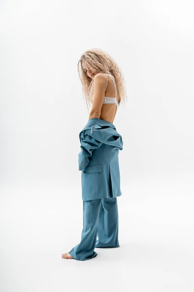 Full length of provocative and barefoot woman in bra and blue oversize suit taking off blazer while standing on grey background, wavy ash blonde hair, slender body, modern individuality — Stock Photo