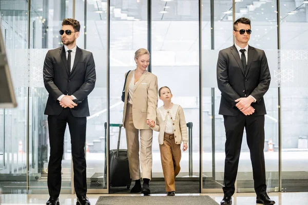 Private security concept, two bodyguards in formal wear and sunglasses standing near hotel entrance, cheerful woman and child holding hands and walking with luggage, entering lobby, luxury lifestyle — Stock Photo
