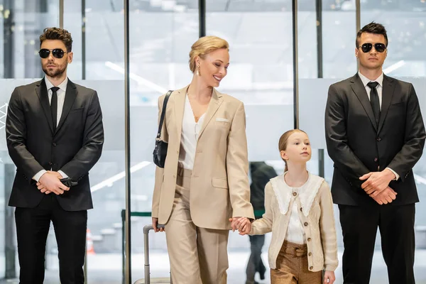 Private security service, two bodyguards in formal wear and sunglasses standing near hotel entrance, happy woman and child holding hands and walking with luggage, entering lobby, luxury lifestyle — Stock Photo