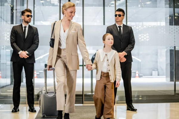 Personal security service, two bodyguards in formal wear and sunglasses standing near hotel entrance, happy mother and child holding hands and walking with luggage, entering lobby, luxury lifestyle — Stock Photo